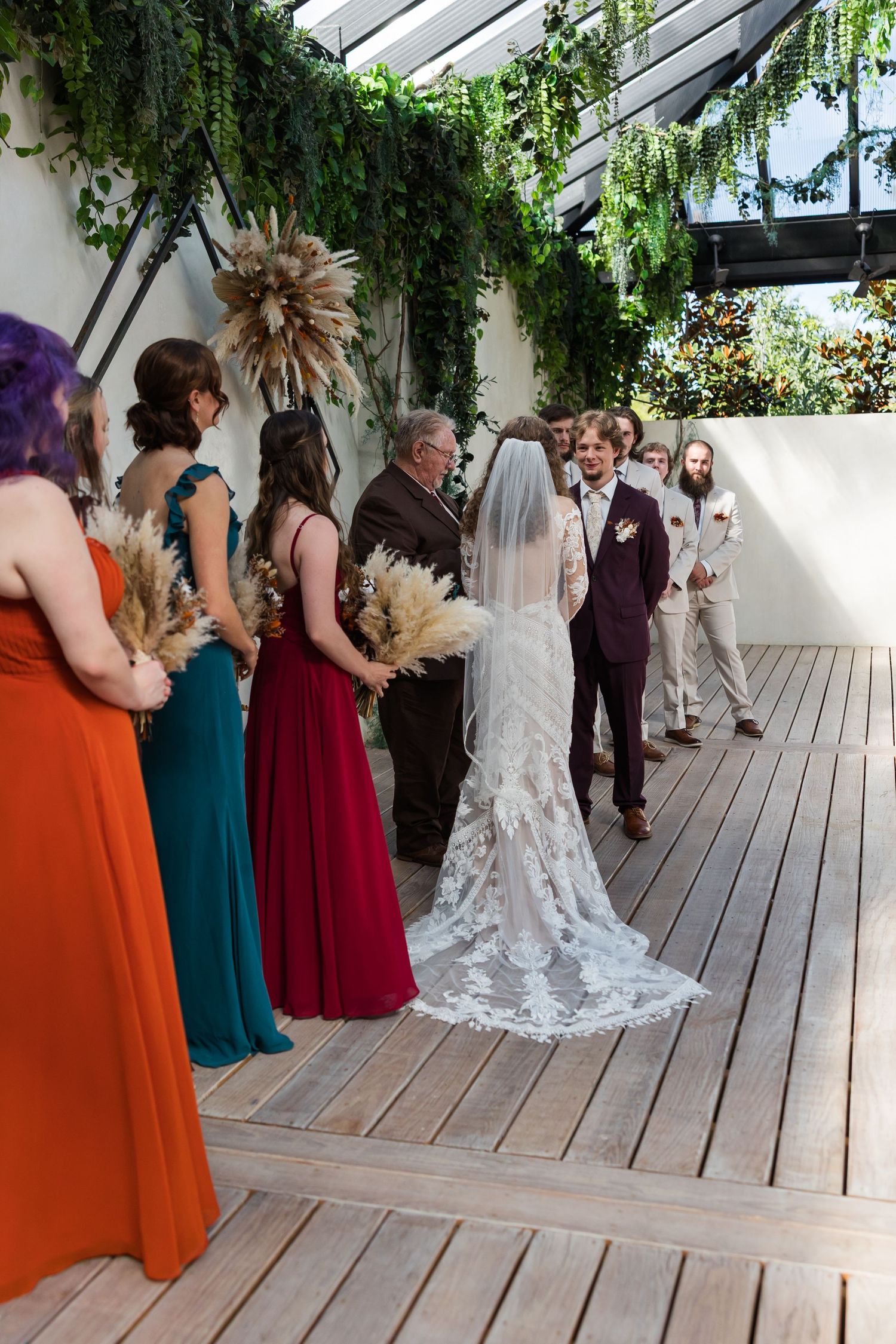 Bride in lace gown with cathedral veil stands at altar with bridesmaids in jewel-toned dresses during ceremony.