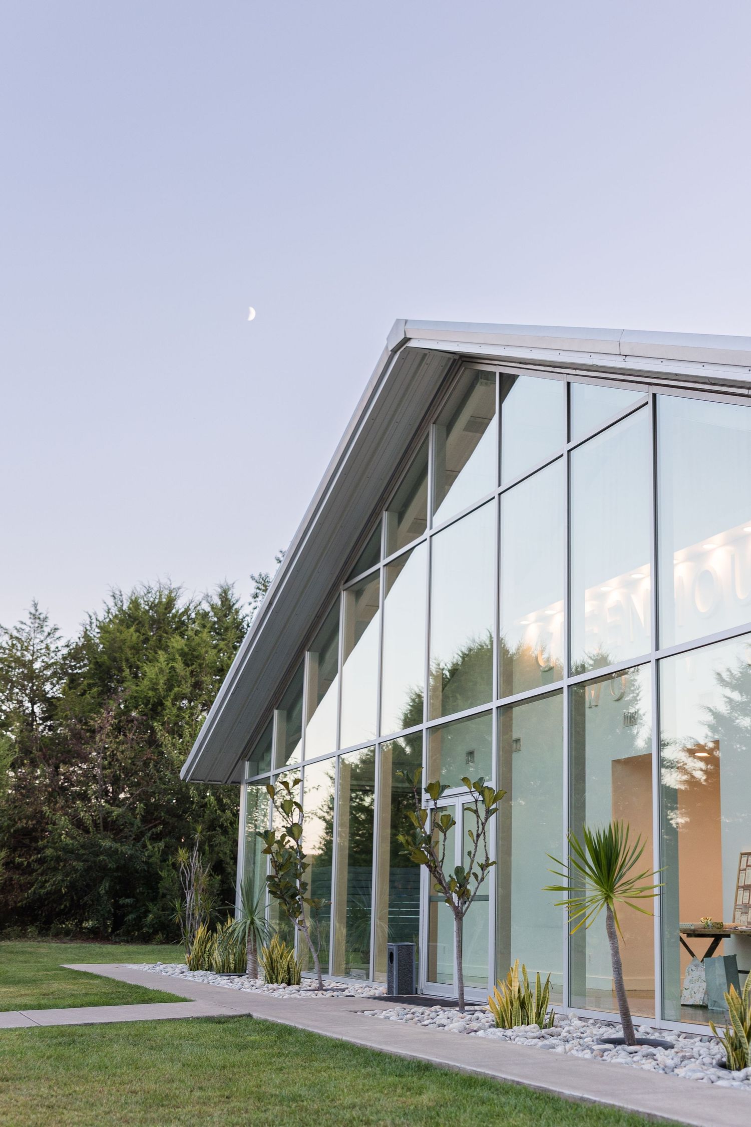 Modern glass building with floor-to-ceiling windows surrounded by lush greenery under a clear sky with a crescent moon.