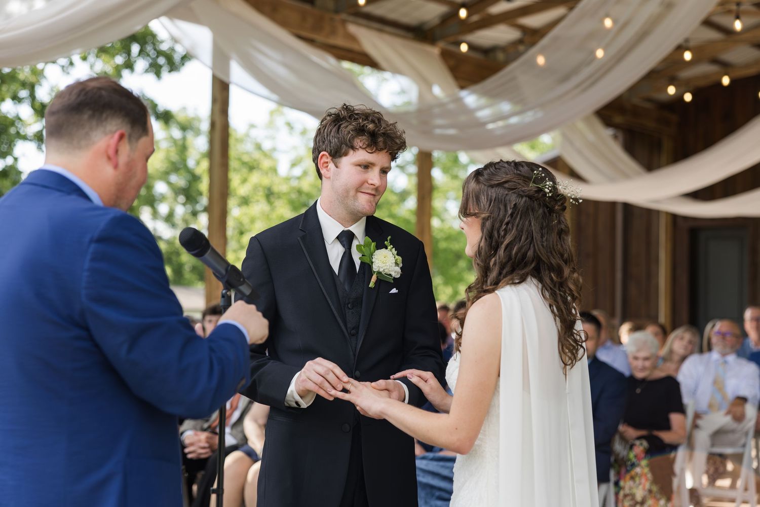 Groom places wedding ring on brides finger during outdoor barn wedding ceremony with officiant.