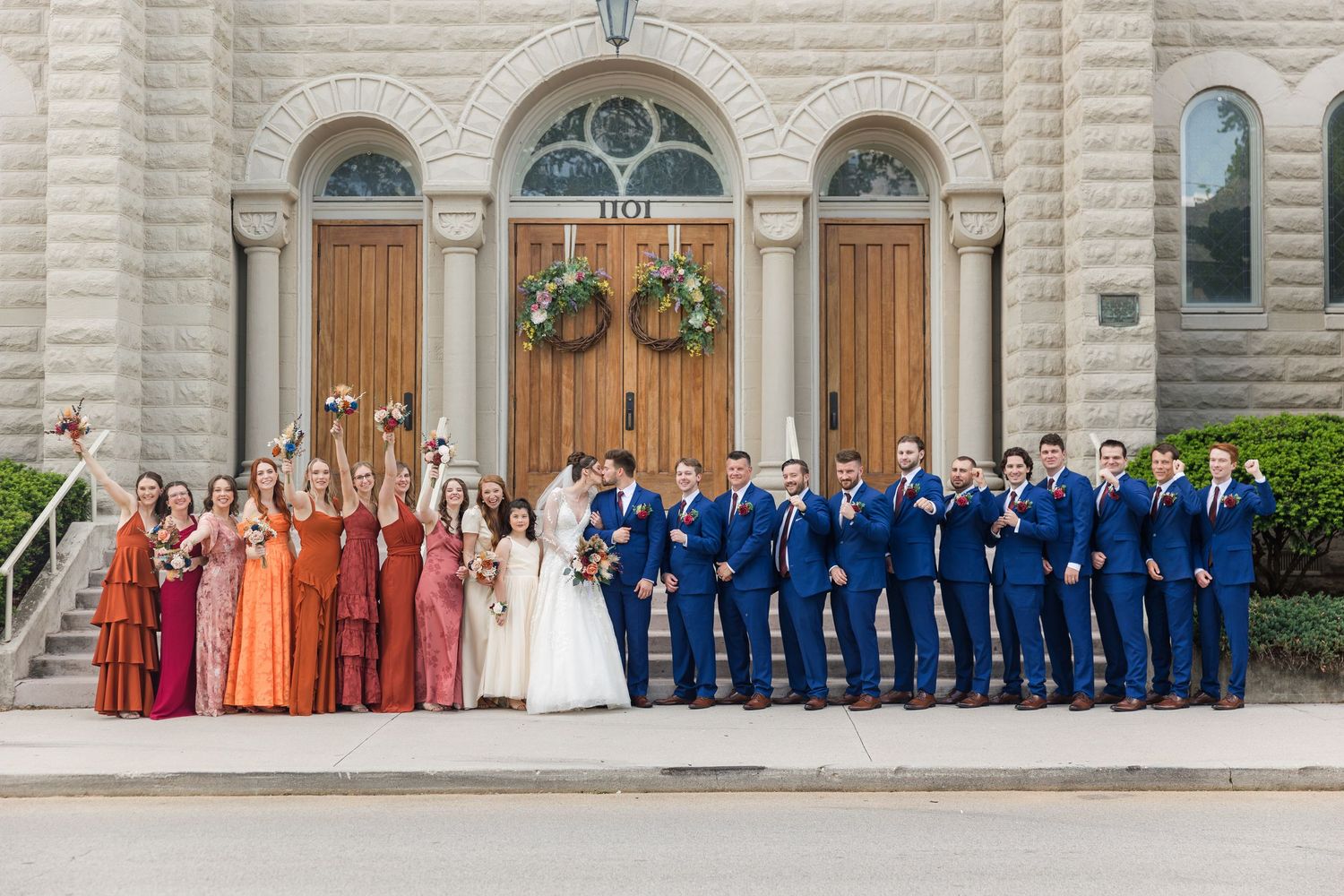 Large wedding party poses before stone church doors, bridesmaids in rust and pink gowns, groomsmen in navy suits.