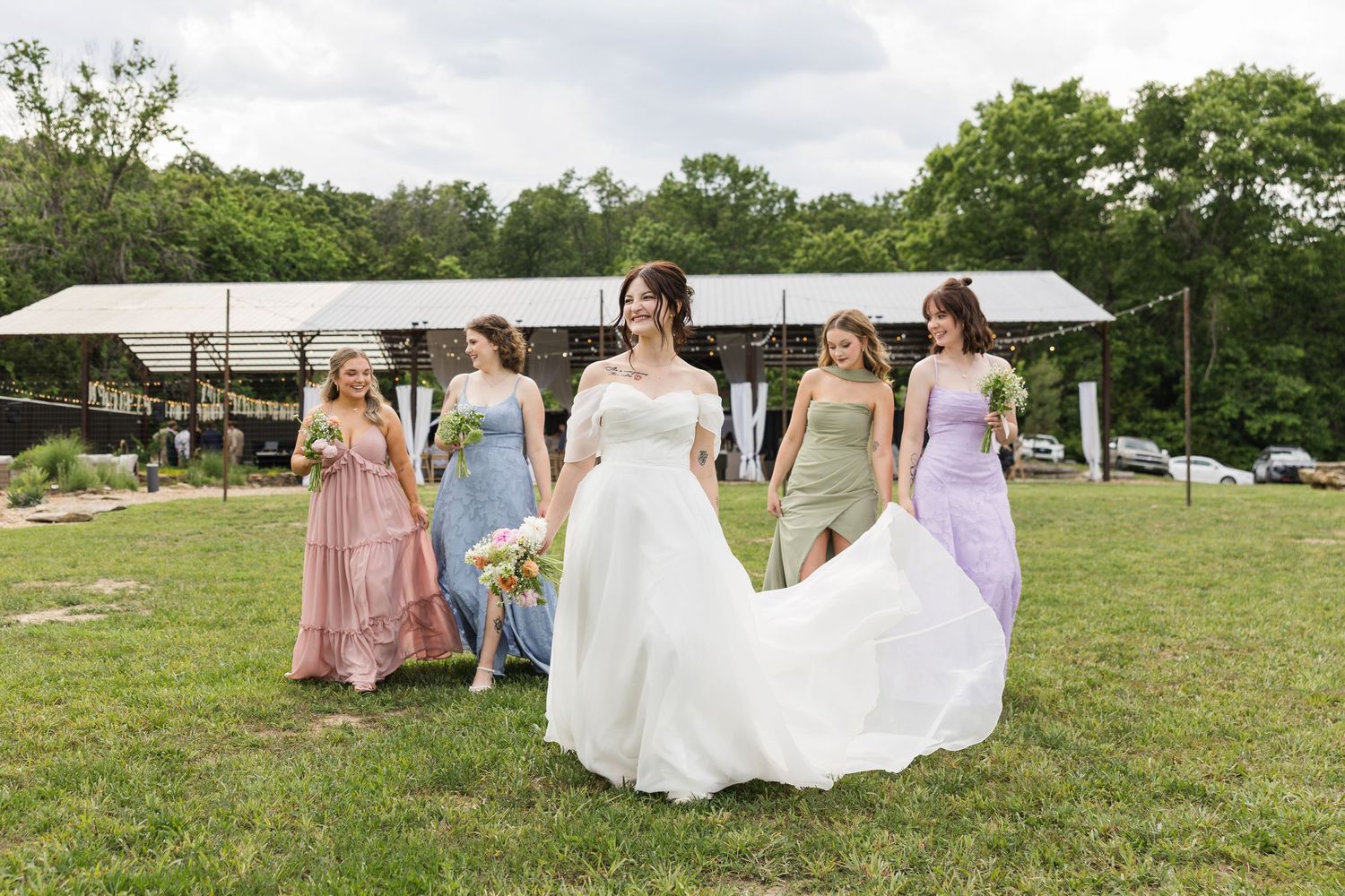 Bride in white gown walks with four bridesmaids in colorful mismatched dresses on green lawn at outdoor wedding venue.