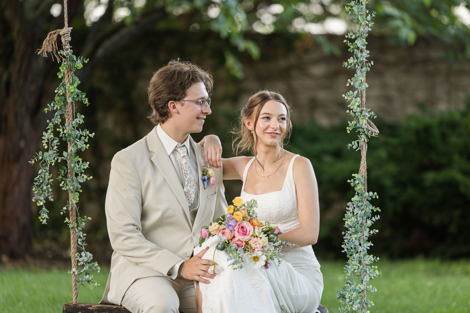 A bride and groom sit together on a garden swing, smiling, as she holds a colorful bouquet on their wedding day.