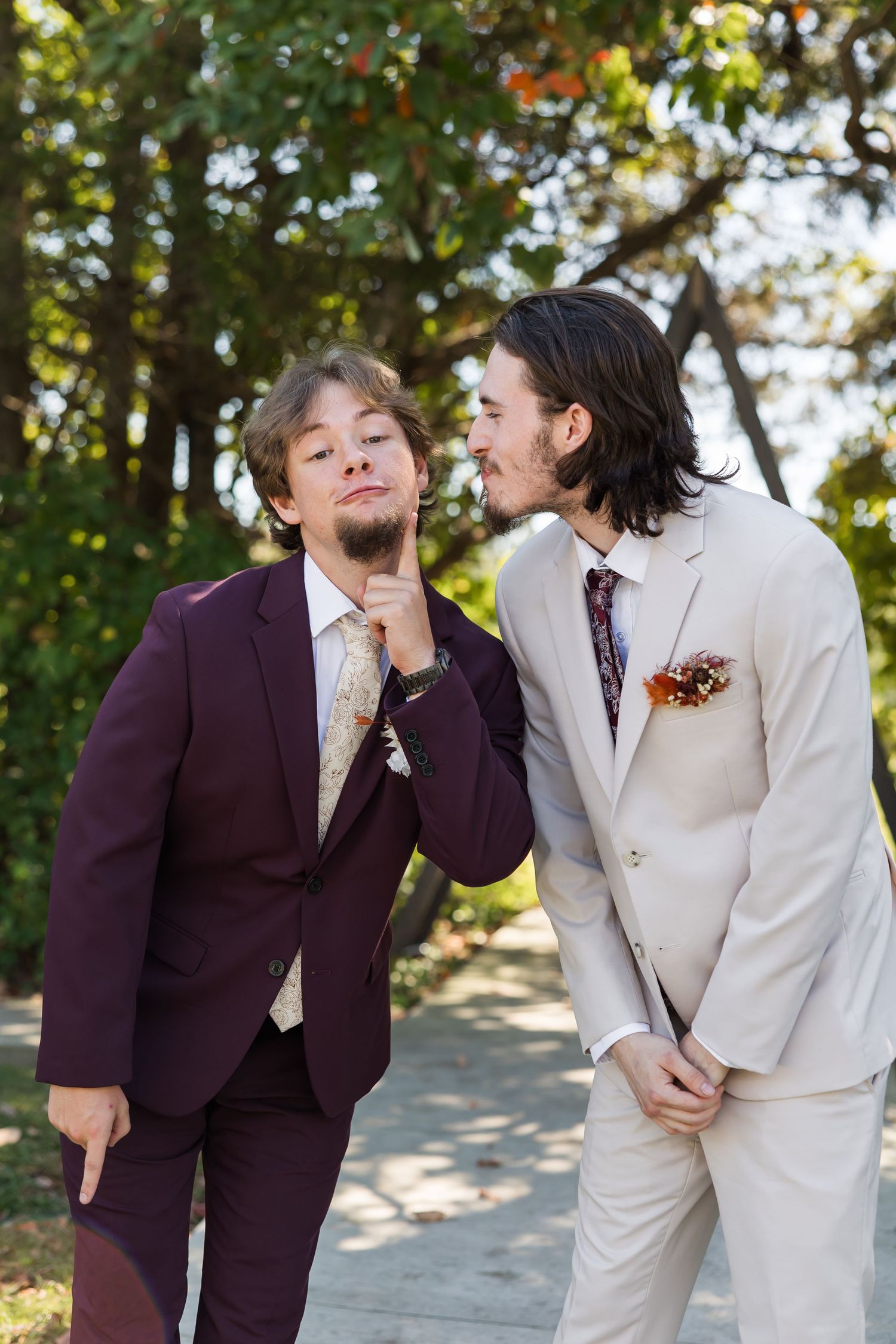 Two men in suits joke around outdoors, one in burgundy grabbing the other's tie playfully.