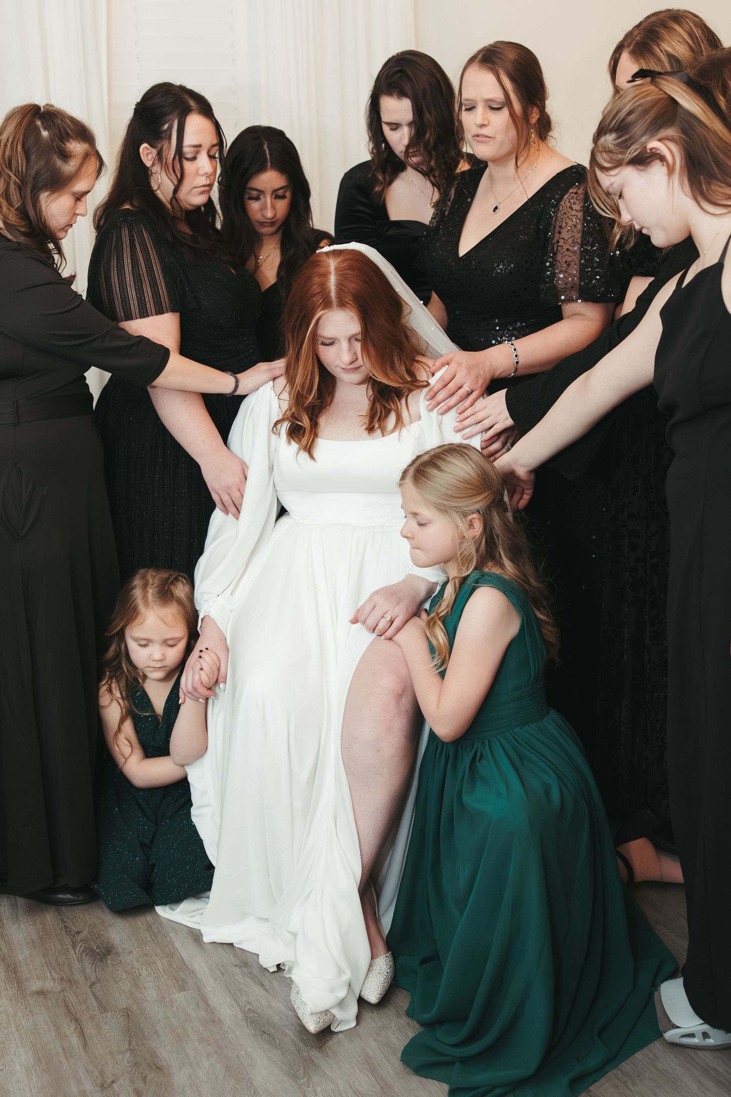 Bride in white gown surrounded by bridesmaids in black dresses during an emotional prayer circle moment.