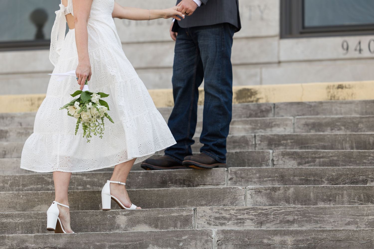 Bride in white dress holding bouquet and groom in jeans holding hands on stone steps at outdoor wedding ceremony.