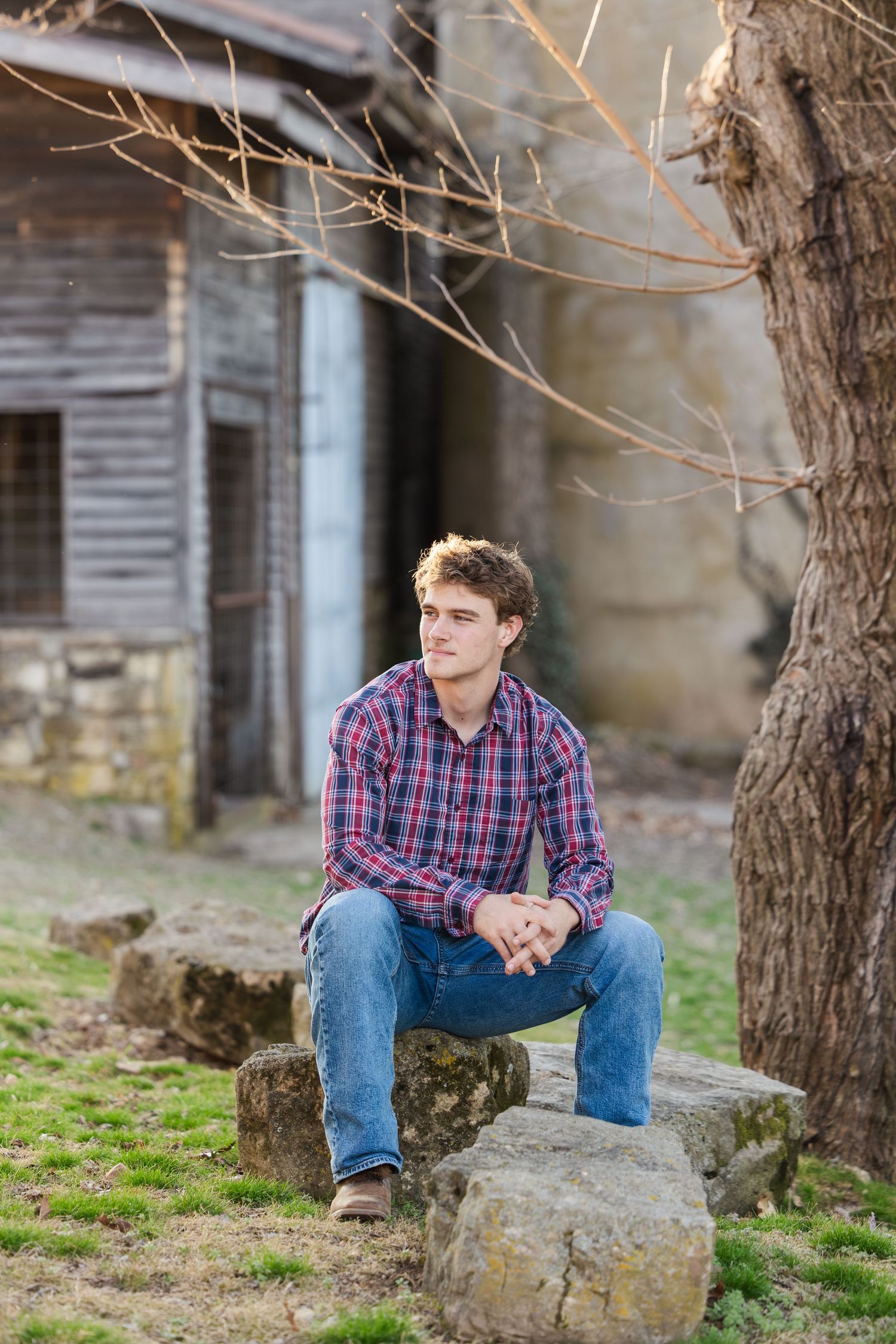 Person in plaid shirt and jeans sitting on rock near rustic building with bare tree branches overhead.