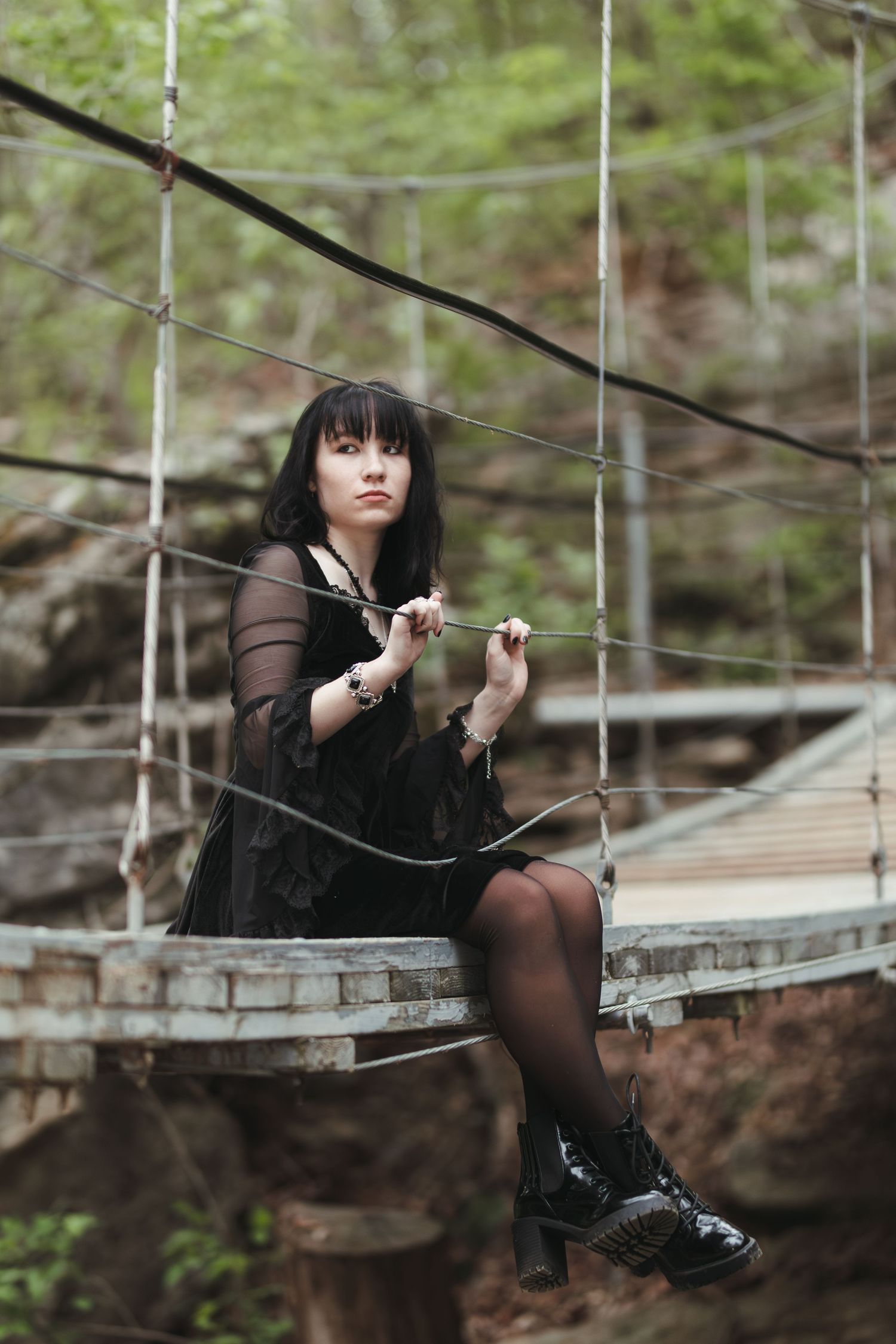 A person in gothic attire sits on a suspended bridge in a moody forest setting, wearing black clothes and combat boots.