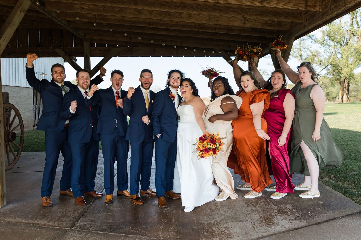 Wedding party wearing navy and colorful dresses poses together under rustic wooden shelter.