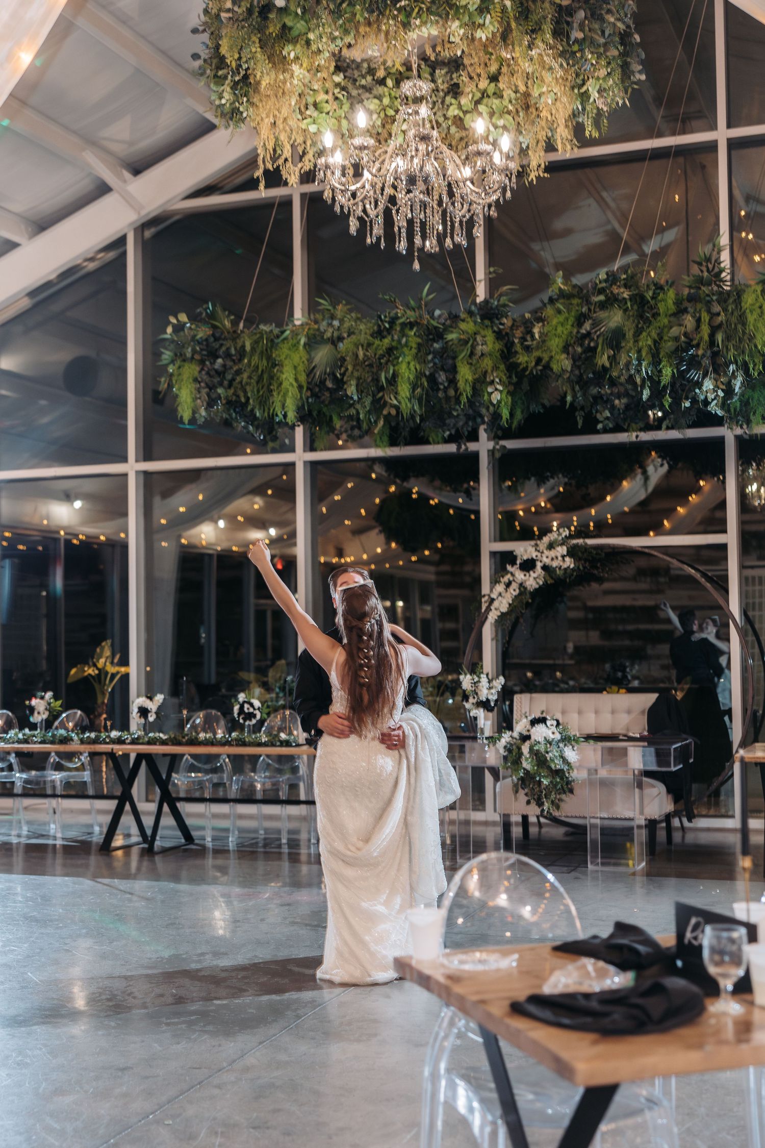 Bride in flowing white dress dancing under crystal chandelier in modern reception venue.