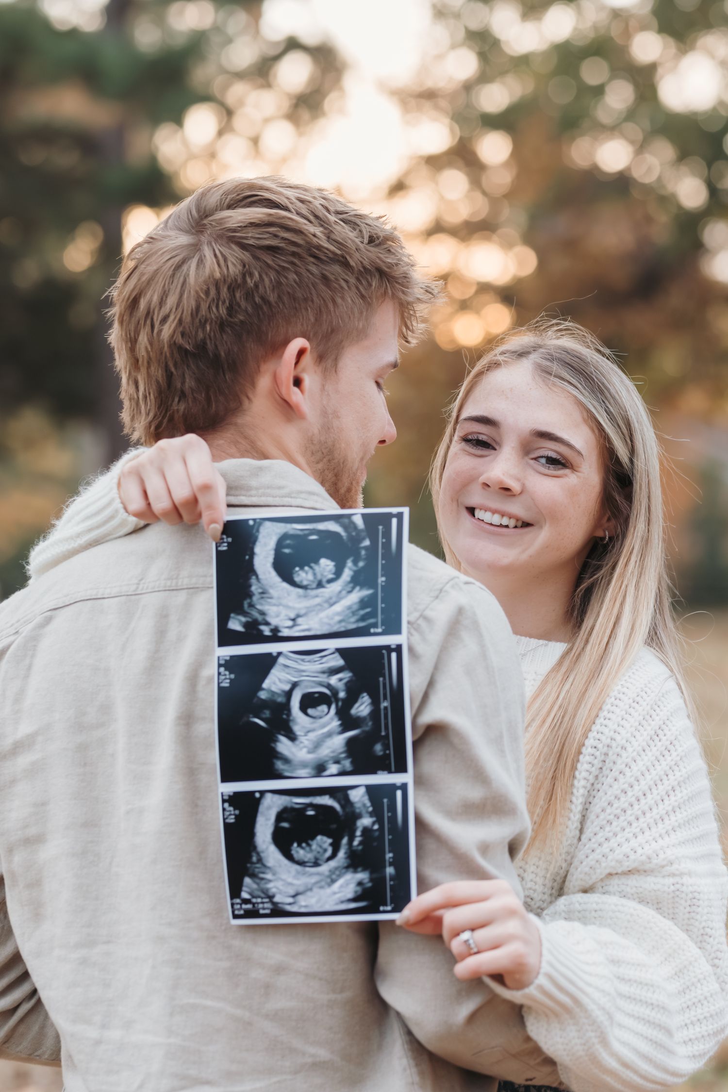 Couple sharing a tender moment while holding ultrasound photos in autumn setting.