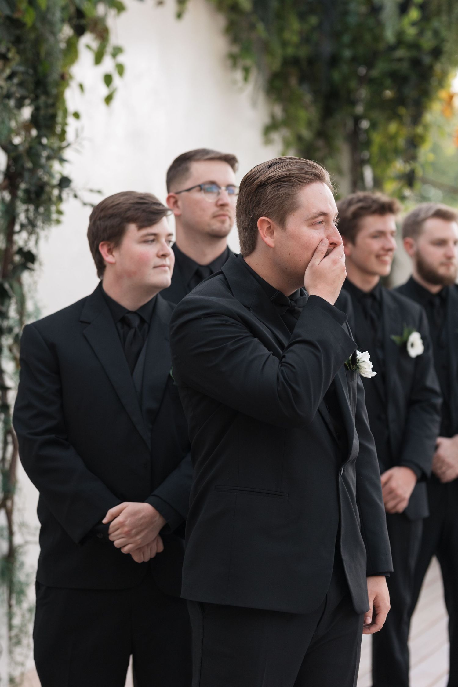 Groomsmen in black suits react emotionally during wedding ceremony.