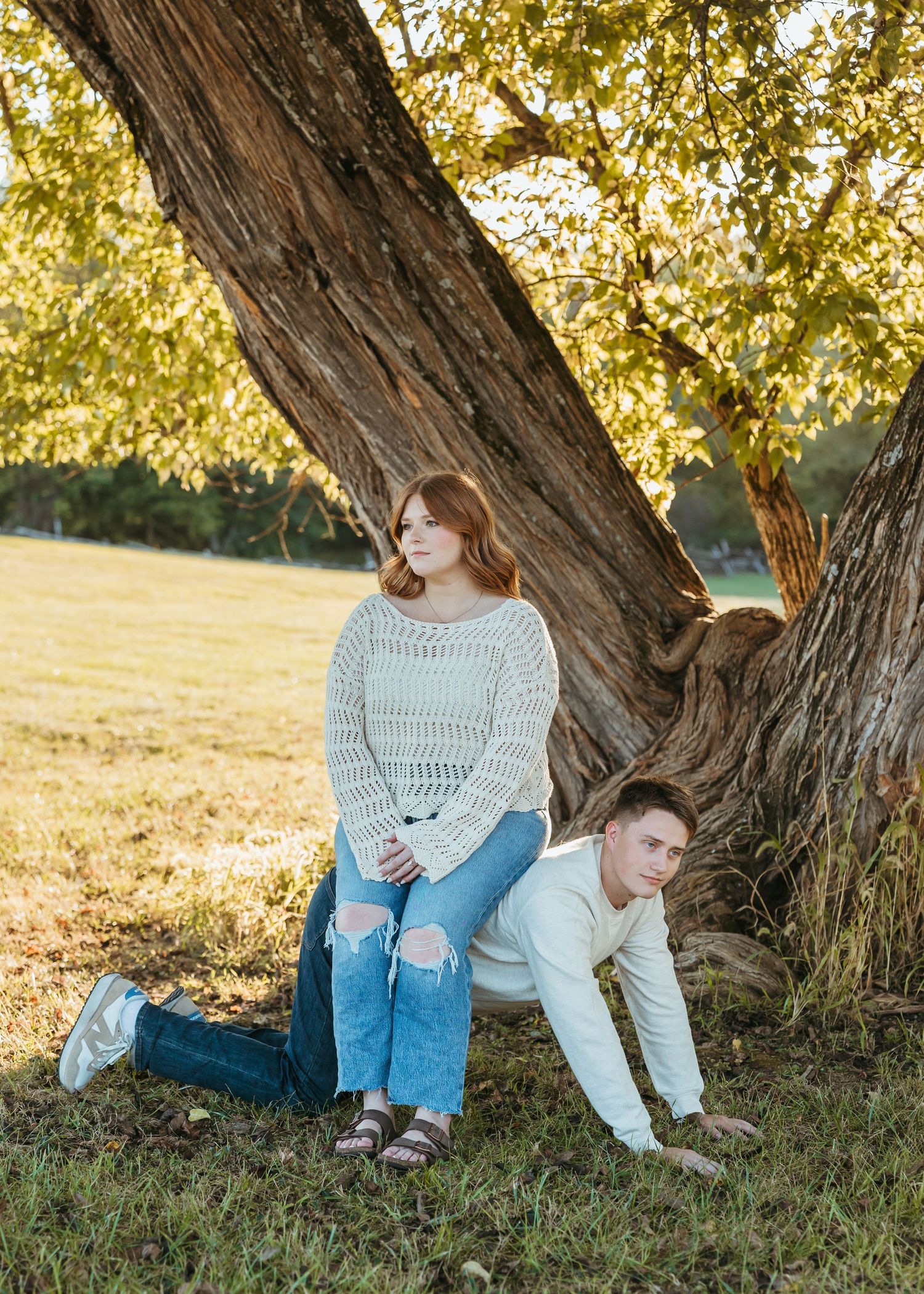 Two people posing playfully by a large tree with autumn leaves in the background.