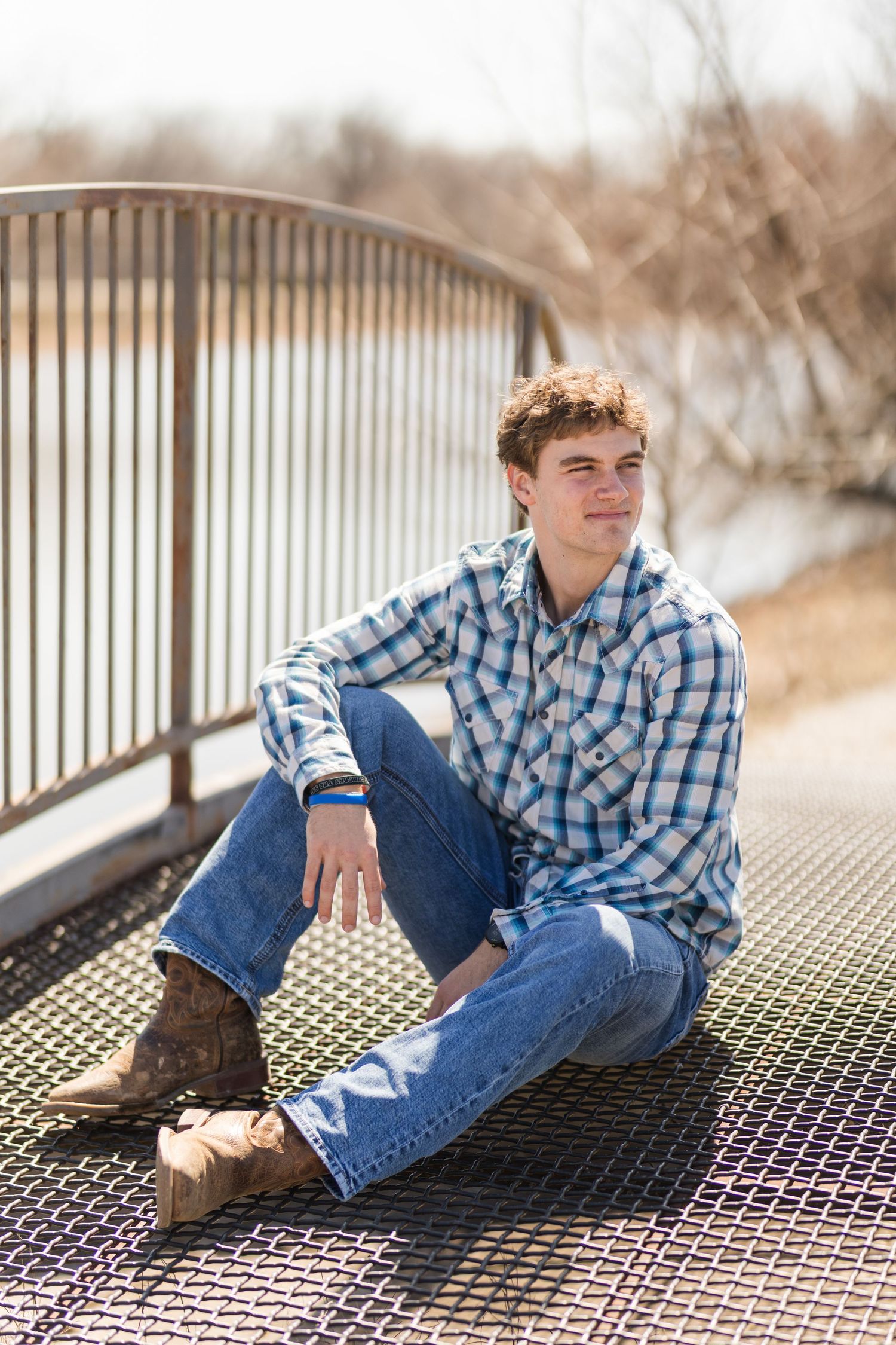 A person in a blue plaid shirt and jeans sits casually on a metal bridge railing on a cloudy day.
