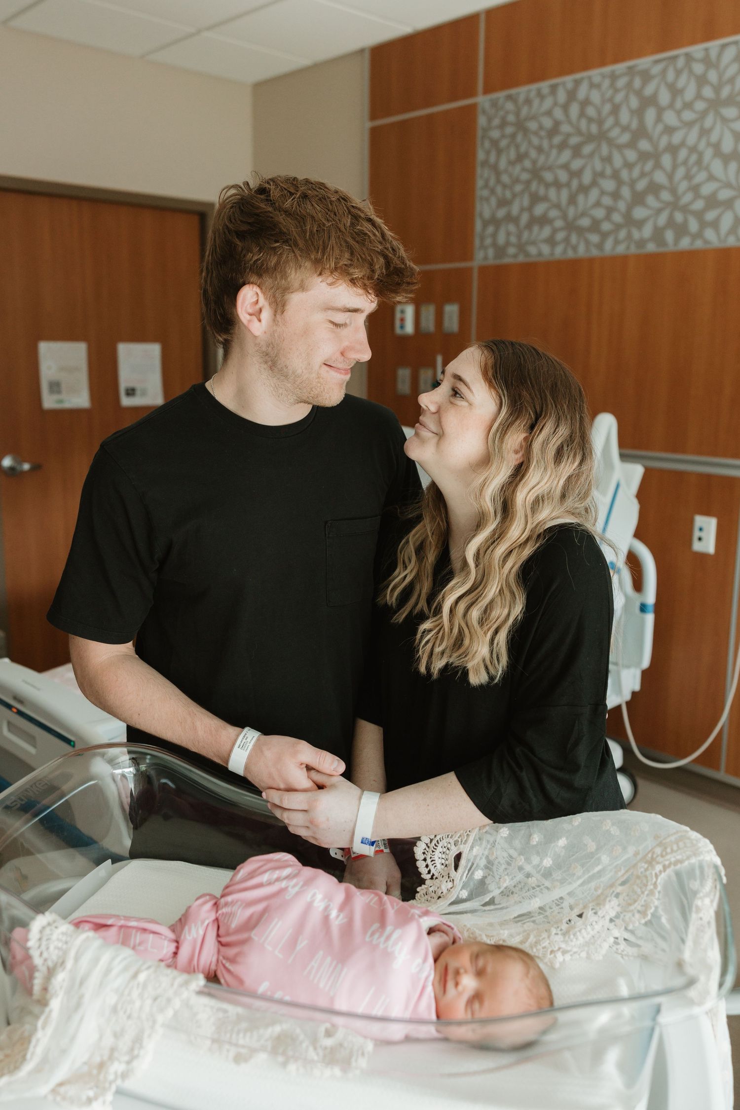 New parents share a tender moment while looking at their newborn baby in a hospital room bassinet.