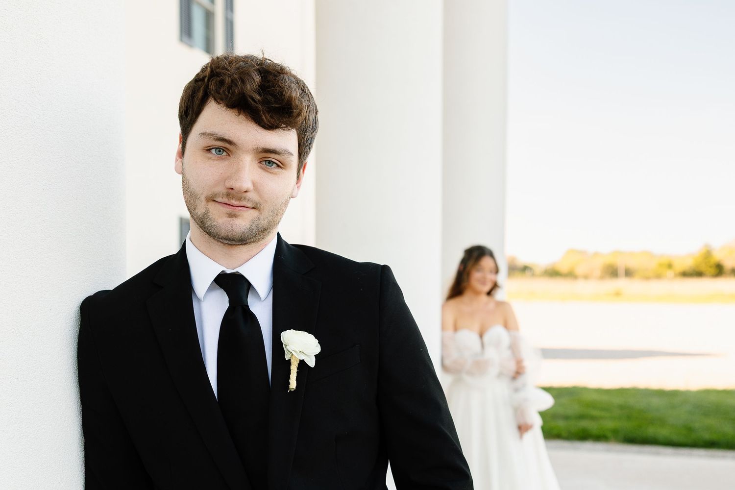 A groomsman in a black suit with a white boutonniere stands indoors near white columns during a formal event.