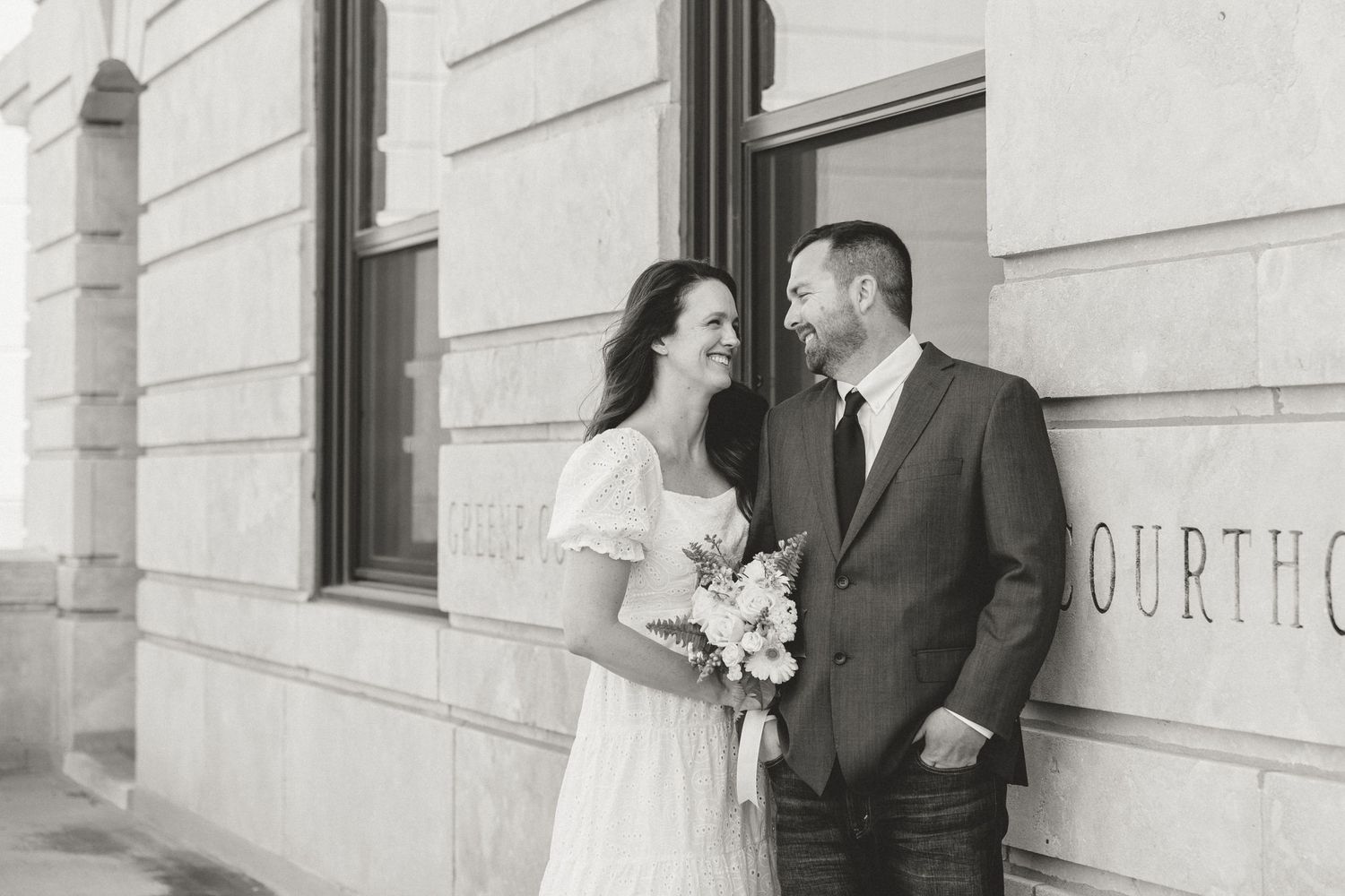 Black and white photo of a smiling bride holding flowers and groom standing outside a courthouse on their wedding day.