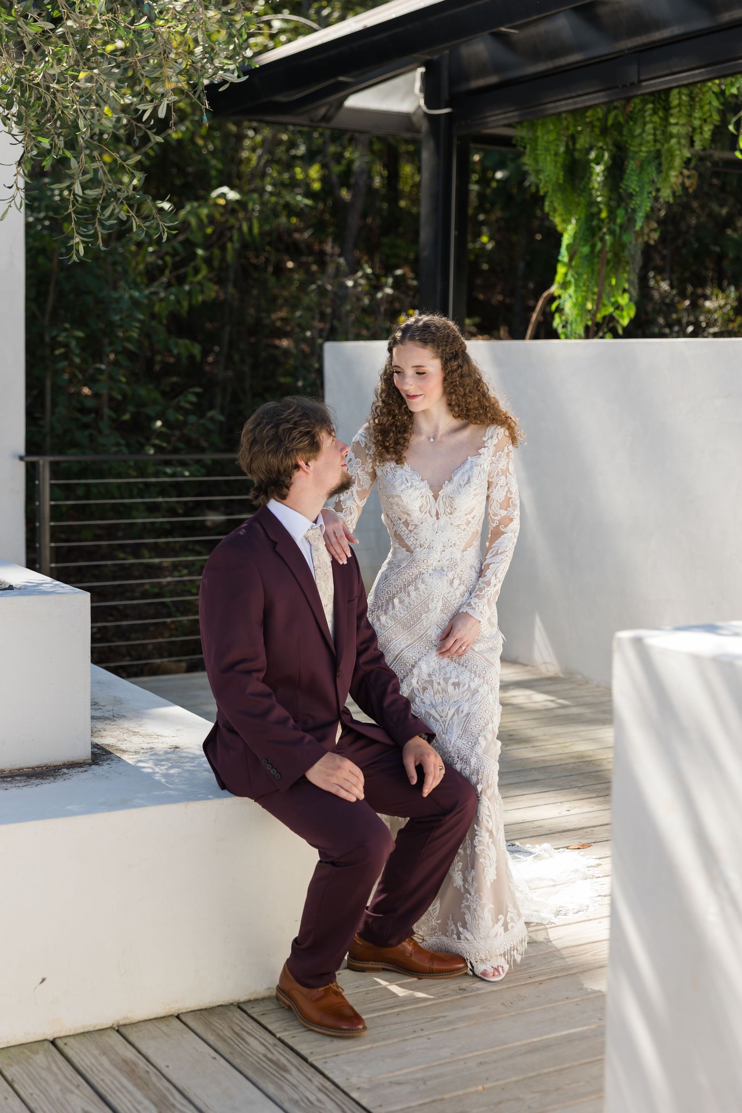 Bride in lace gown and groom in burgundy suit pose together on sunlit outdoor wedding venue deck.