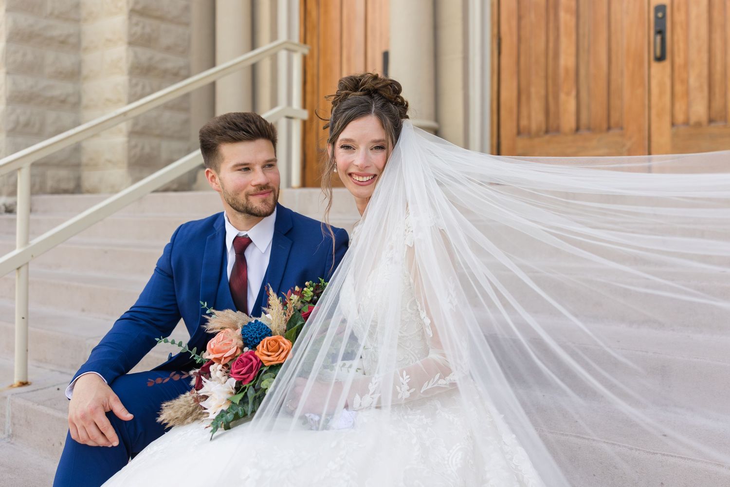 Smiling bride and groom pose on church steps with flowing veil and colorful bouquet.