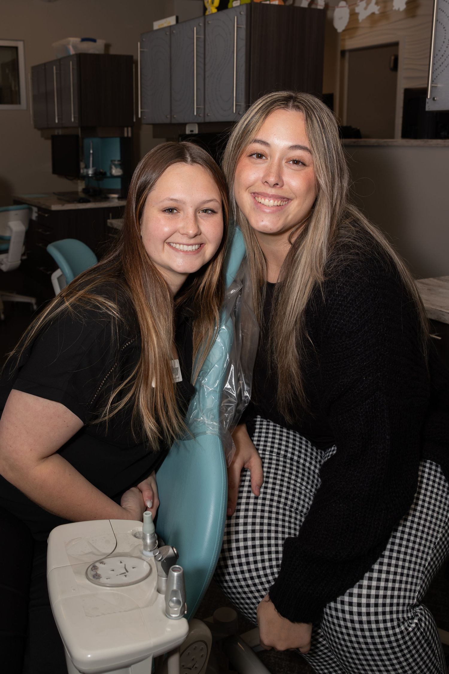 Two women in black tops smile together in a dental office setting.