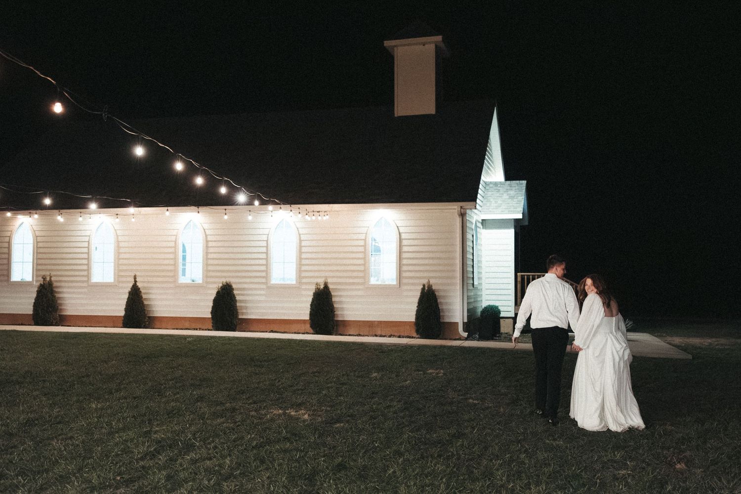 Couple walking outside illuminated white church building at night under string lights.