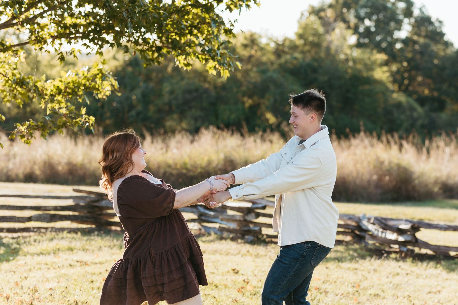 Two people hold hands and dance in a rustic outdoor setting with fall foliage and wooden fencing.