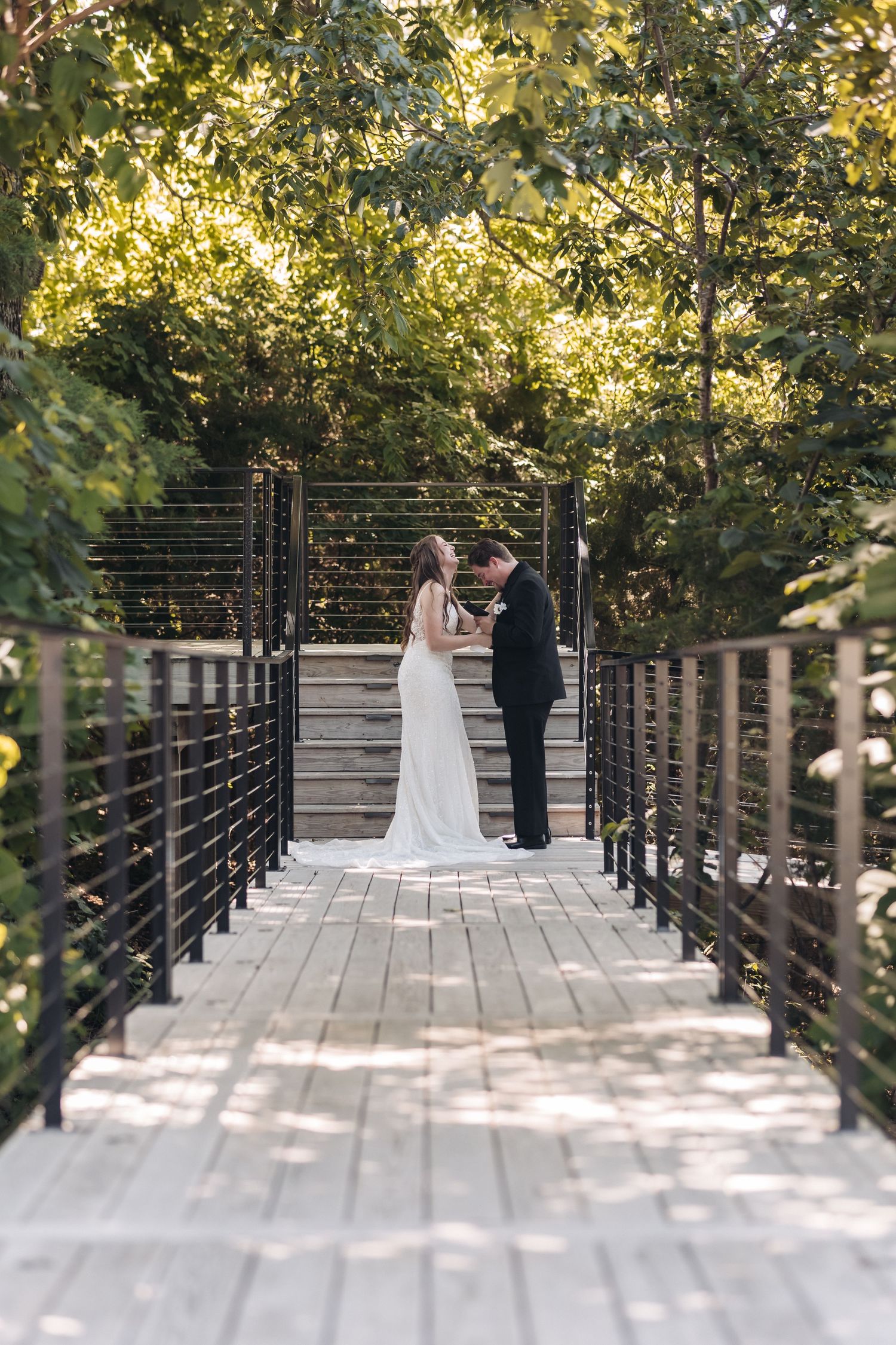 Couple standing on wooden bridge surrounded by lush green trees during wedding ceremony.
