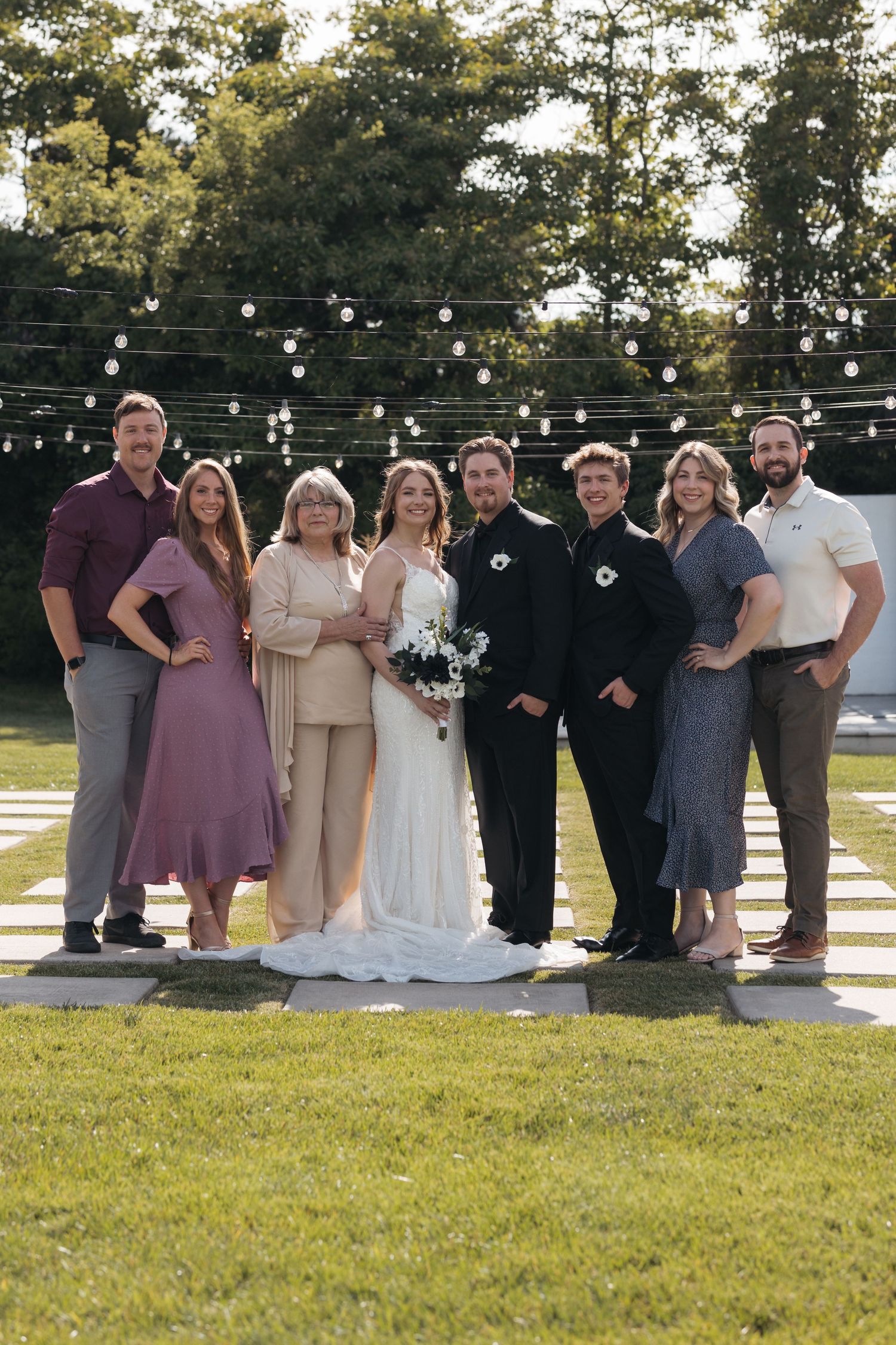 Wedding party poses on a garden path under string lights with greenery in the background.