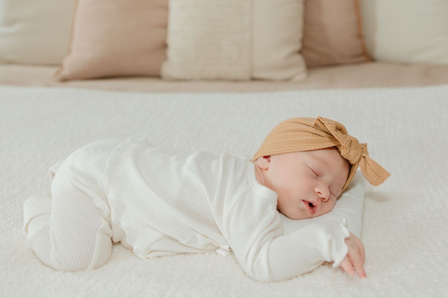 Sleeping newborn in white onesie and peach cap rests peacefully on cream bedding.