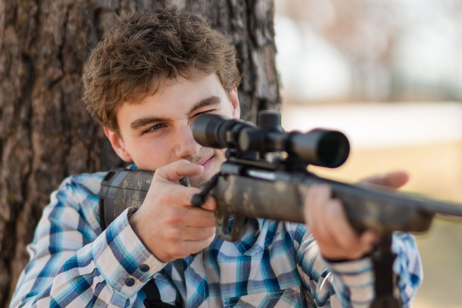 Person in plaid shirt aiming rifle with scope while leaning against tree trunk.