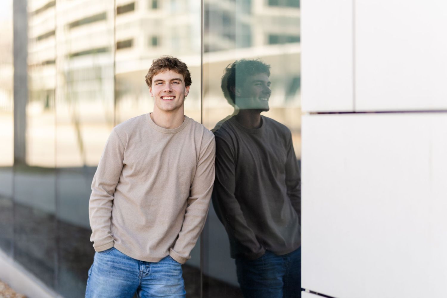 A casual portrait of someone in a beige sweater and jeans standing against a modern building with their reflection visible.