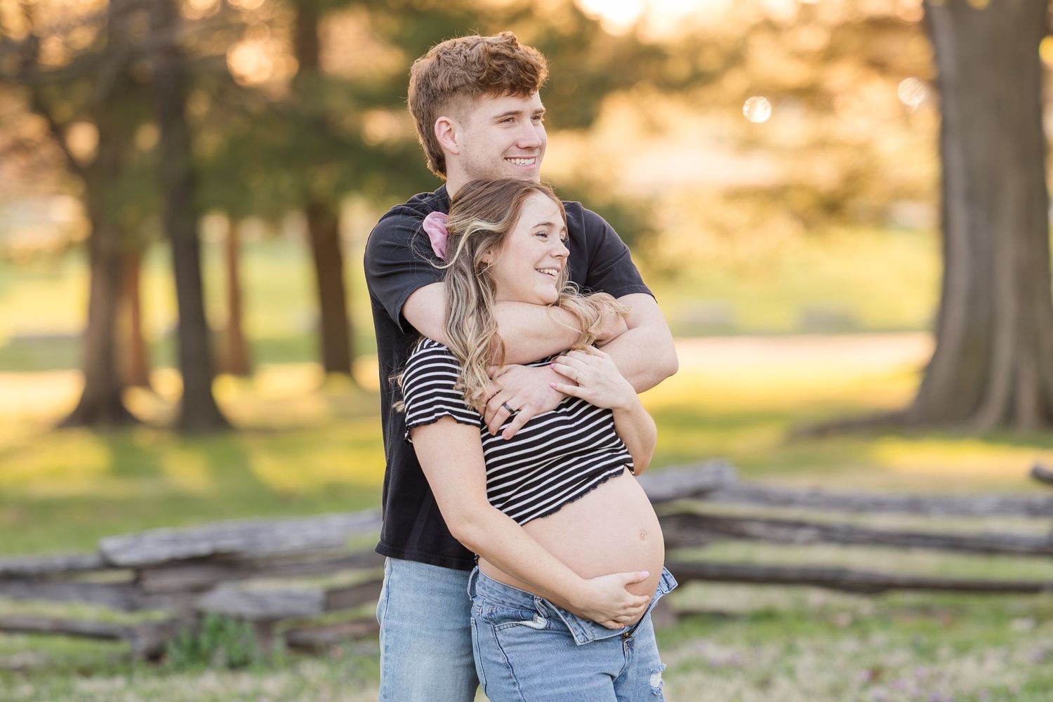 Couple wearing matching striped tops embrace during sunset maternity photoshoot in a park setting.