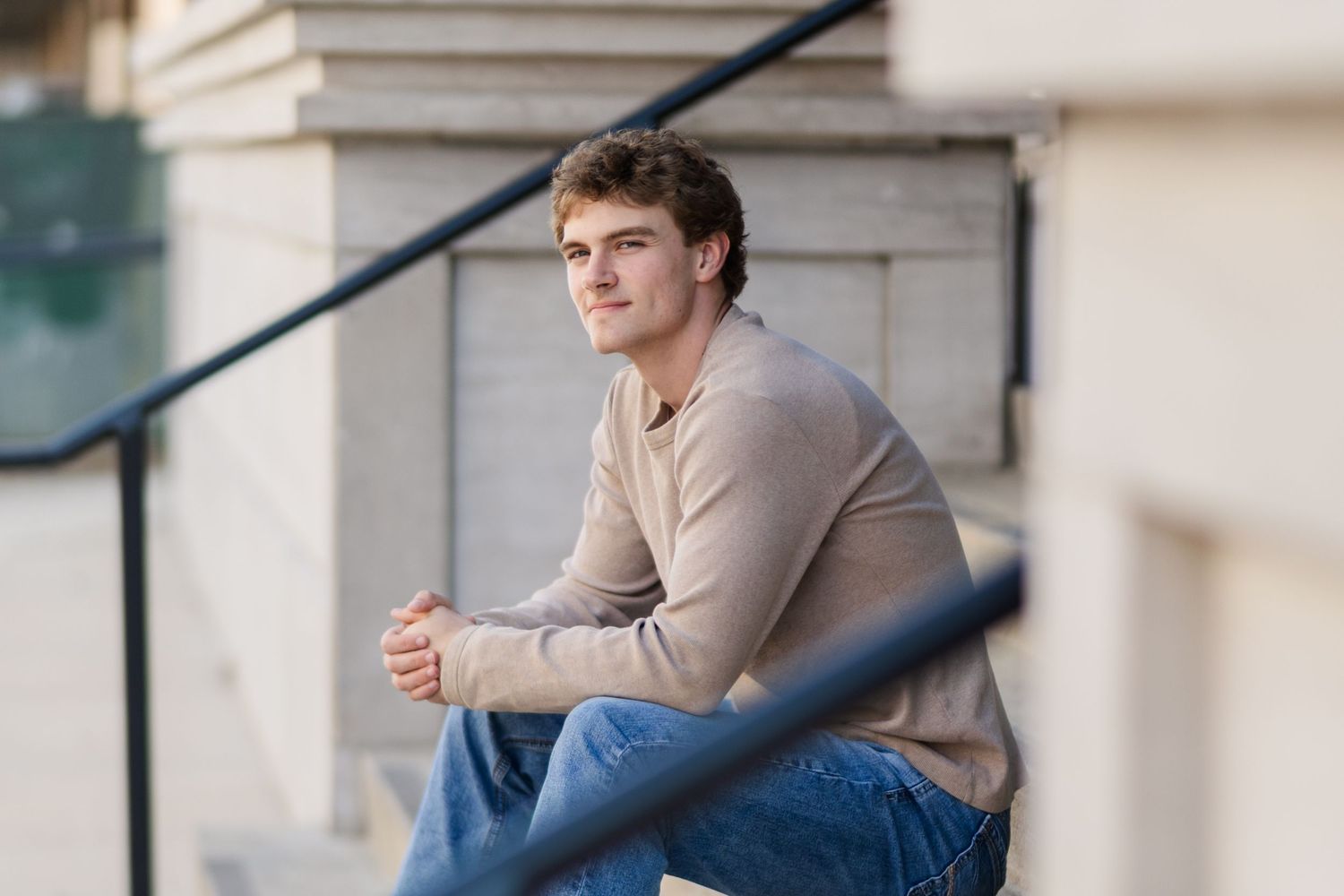 Person in beige sweater and blue jeans sitting on outdoor steps in casual pose.