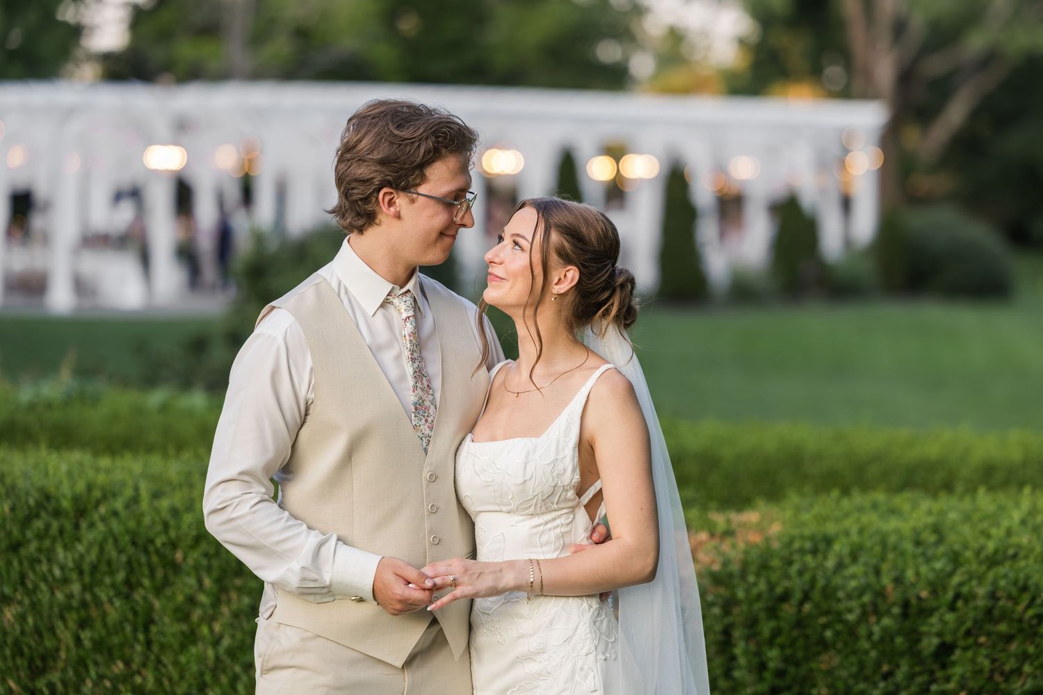 A bride and groom share a romantic moment outdoors, holding hands near lush hedges with a white tent venue behind them.