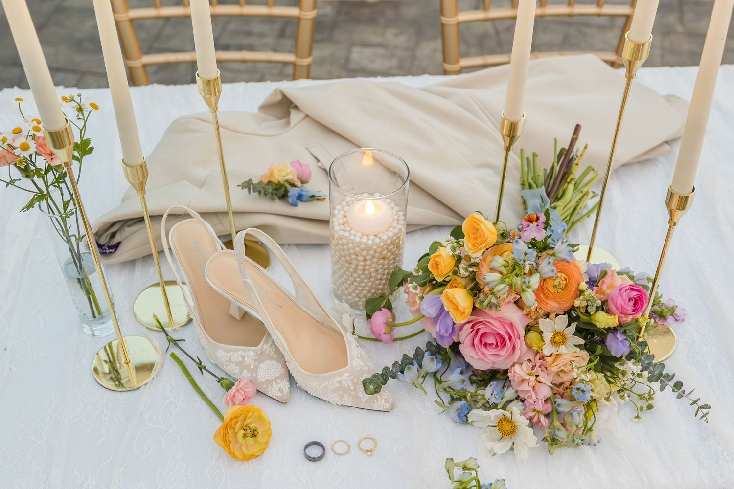 Elegant wedding flat lay with bridal heels, colorful floral bouquet, gold rings, pearl candle, and taper candles on white table.