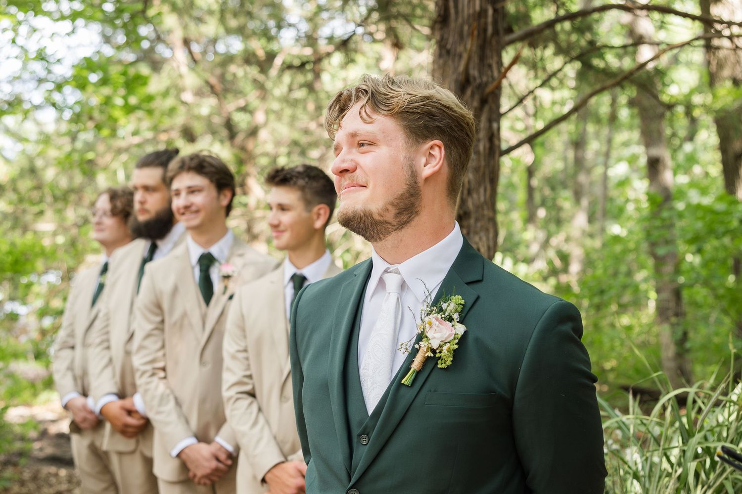 Groom in green suit with boutonniere stands before groomsmen in tan suits at an outdoor forest wedding ceremony.