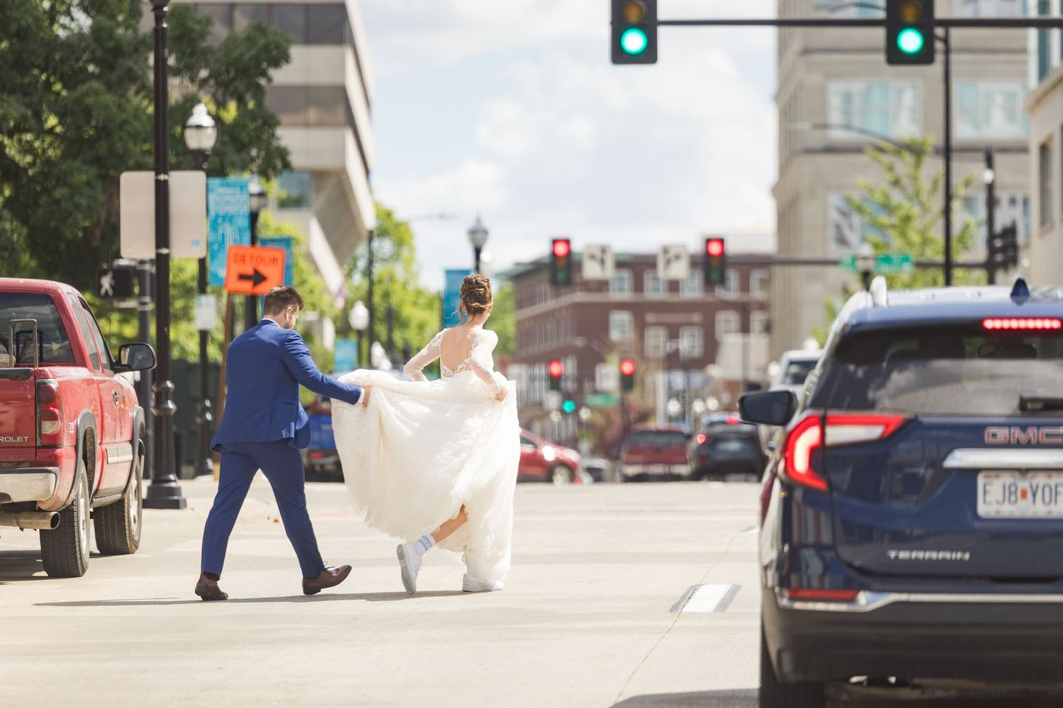 A bride and groom cross a busy city street, the bride holding her white gown as traffic waits at a red light.