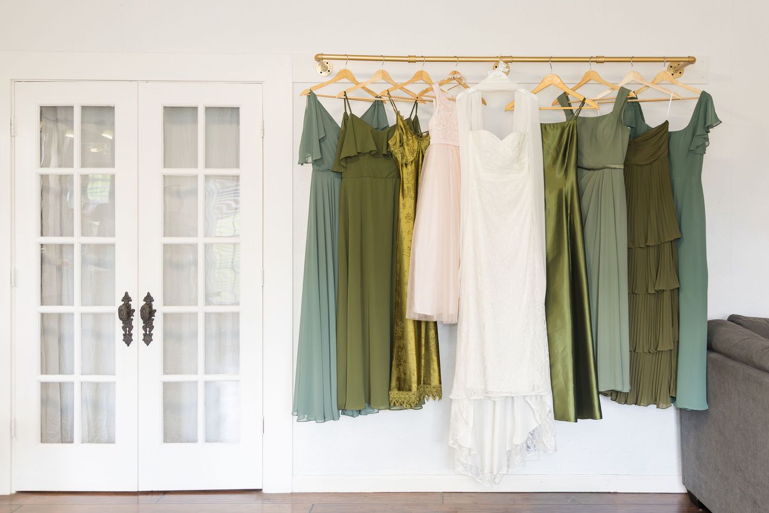Wedding dress and green bridesmaid dresses hanging on a gold rack near white French doors in a bright room.