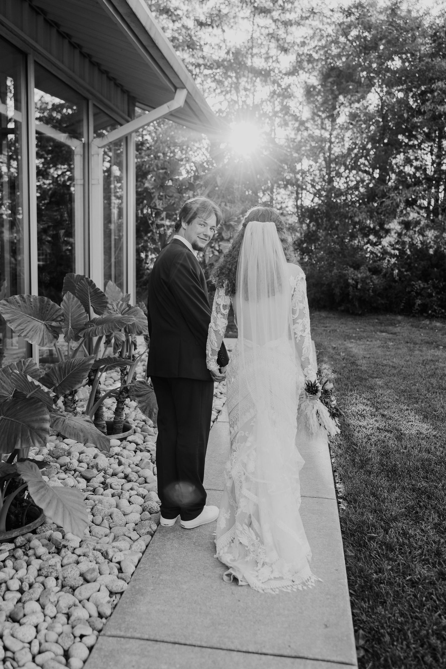 Black and white photo of bride and groom holding hands on a sunlit garden pathway.