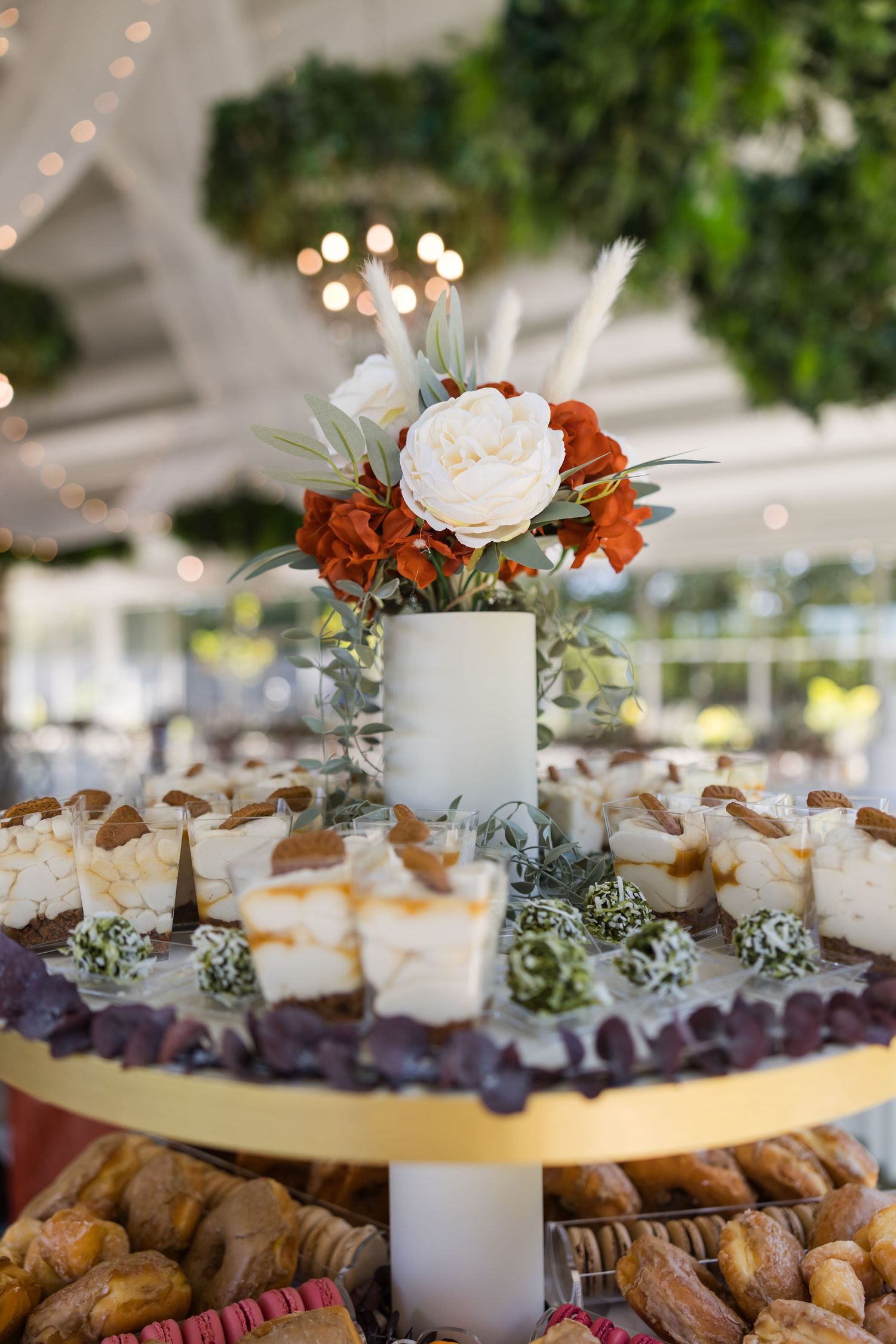 Elegant wedding dessert table with tiered display of pastries, chocolates, and sweets topped with a white and rust floral arrangement.