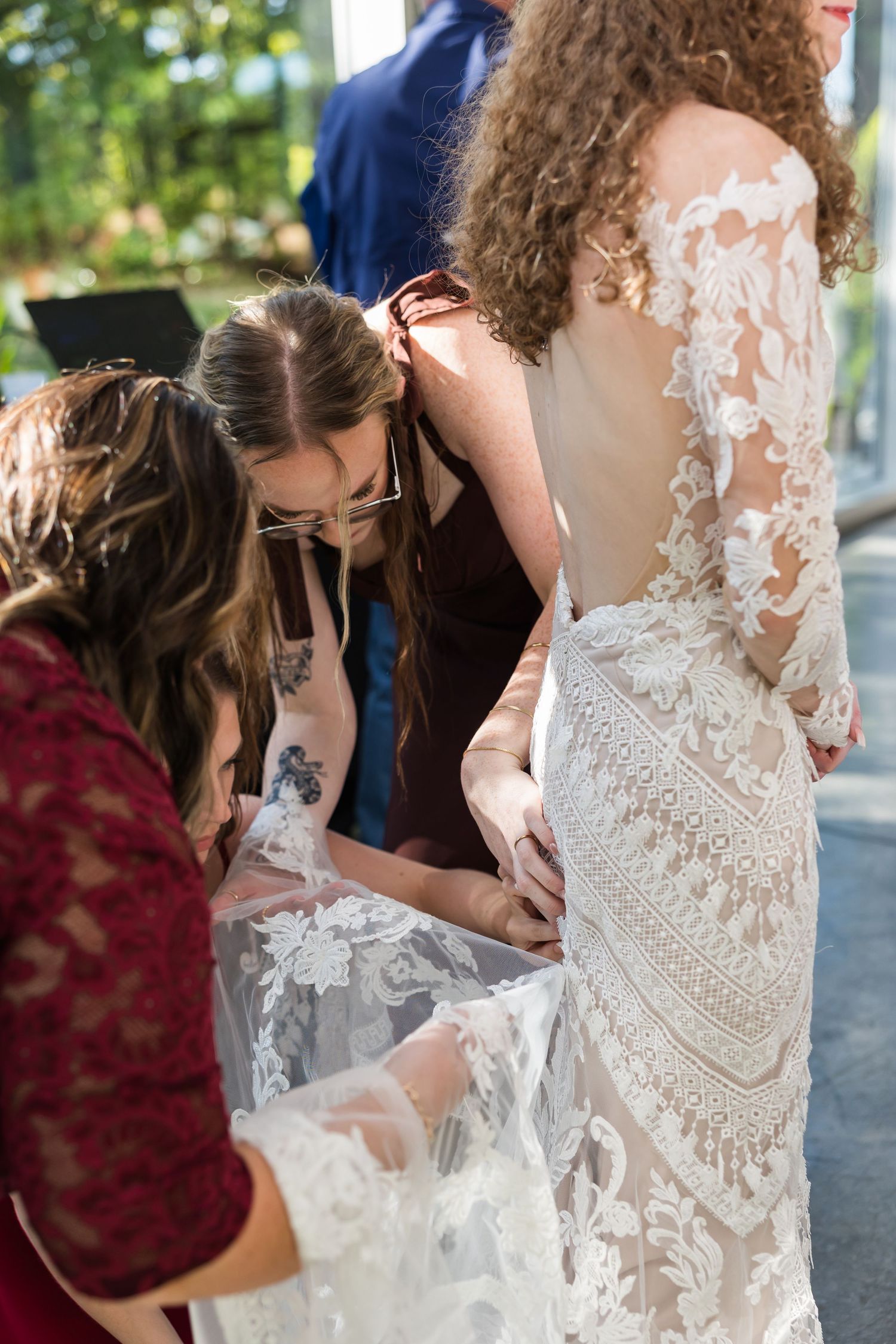 Bridesmaids helping bride in intricate lace backless wedding gown with her dress details before the ceremony.