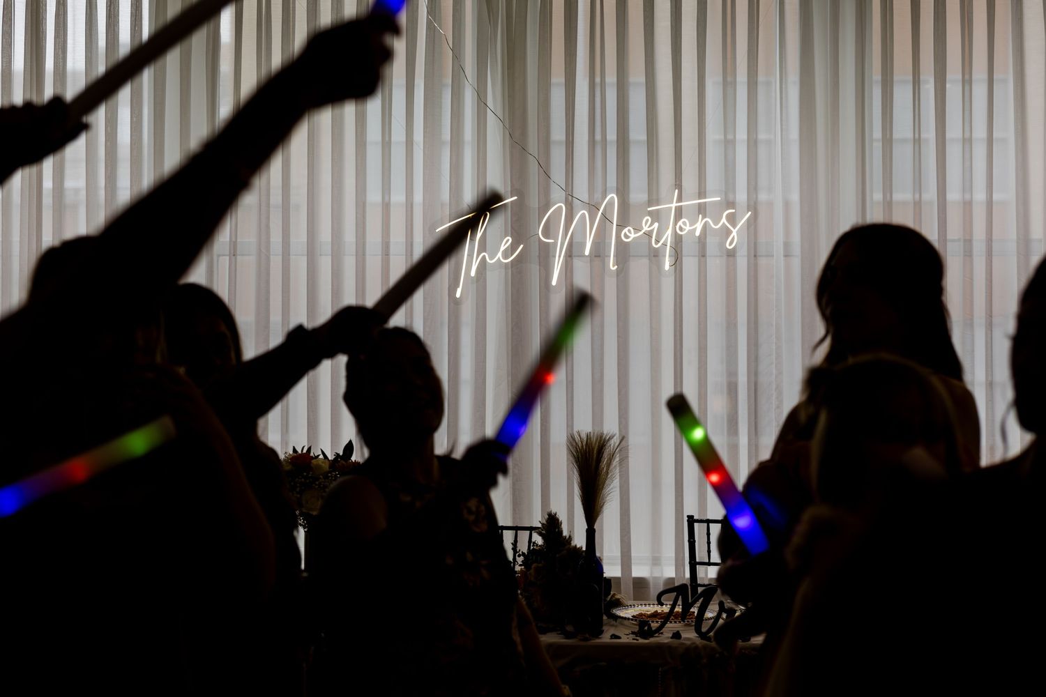 Silhouetted wedding guests wave glowing light sticks under The Mortons neon sign at reception.