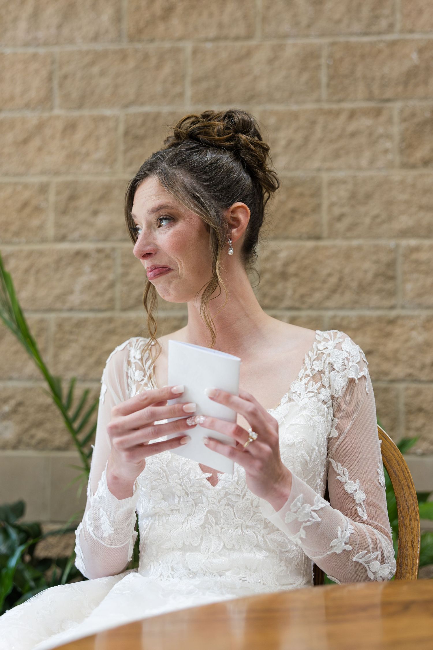 Bride in lace long-sleeve wedding gown holds a card while sitting at a table, gazing thoughtfully to the side.