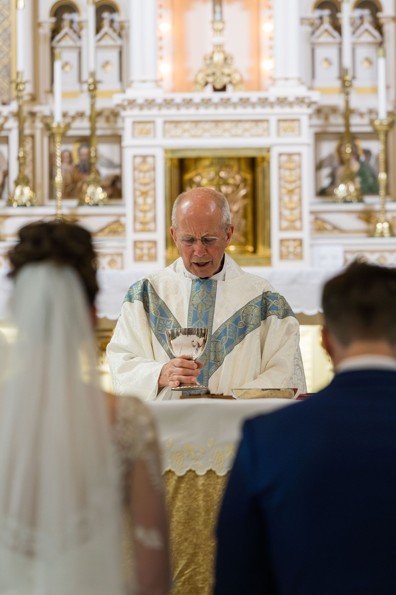 A Catholic priest holds a chalice while officiating a wedding ceremony at an ornate golden altar.