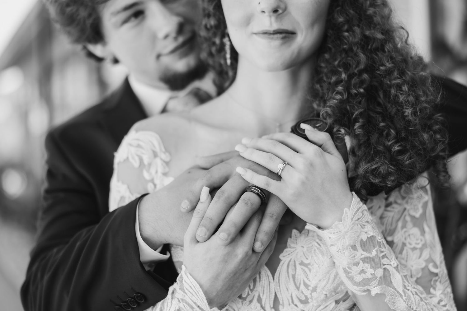 Black and white wedding photo of bride and groom holding hands, showcasing wedding ring, in elegant lace dress.