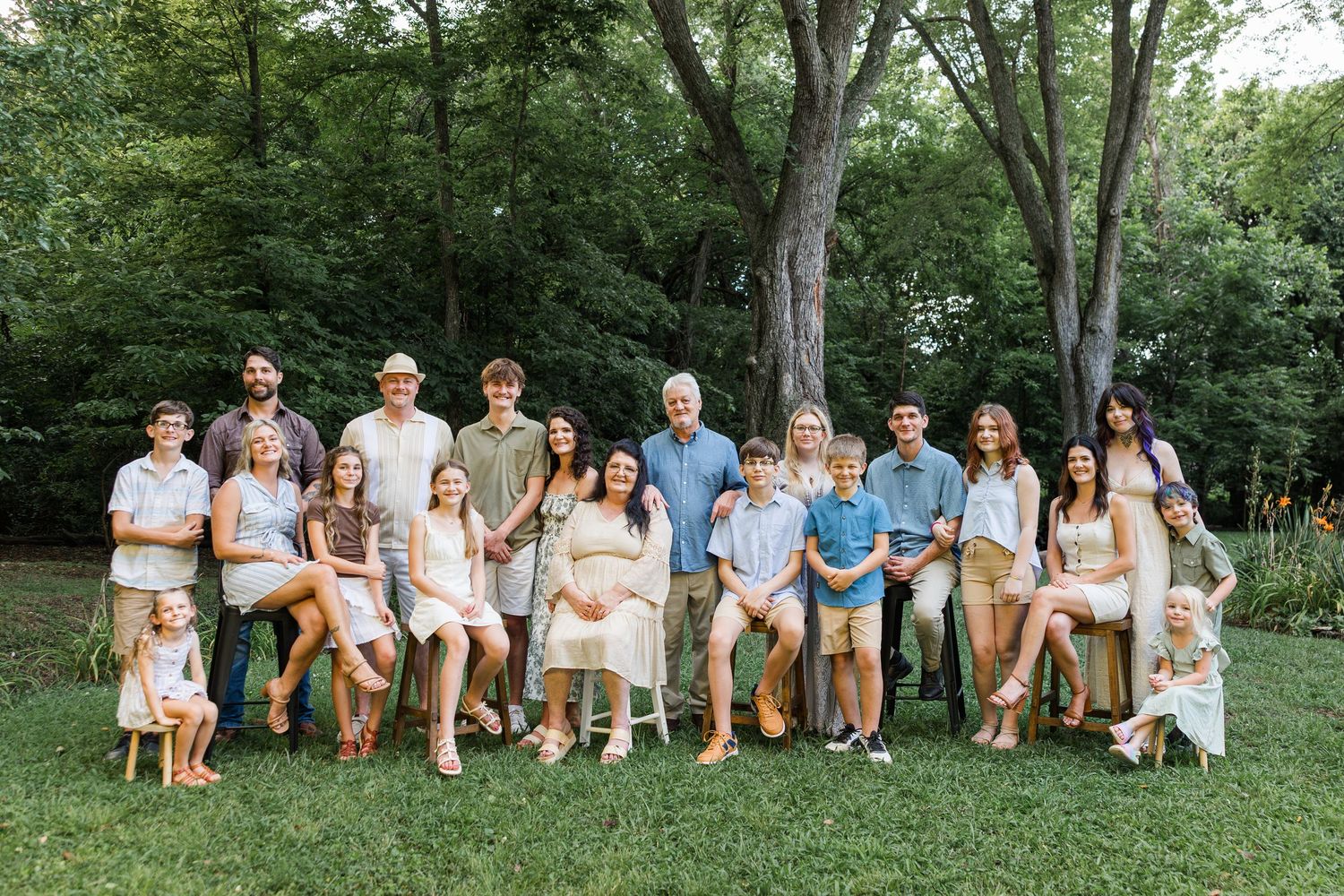 A large group dressed in casual summer attire sits together on benches in a lush green outdoor setting.