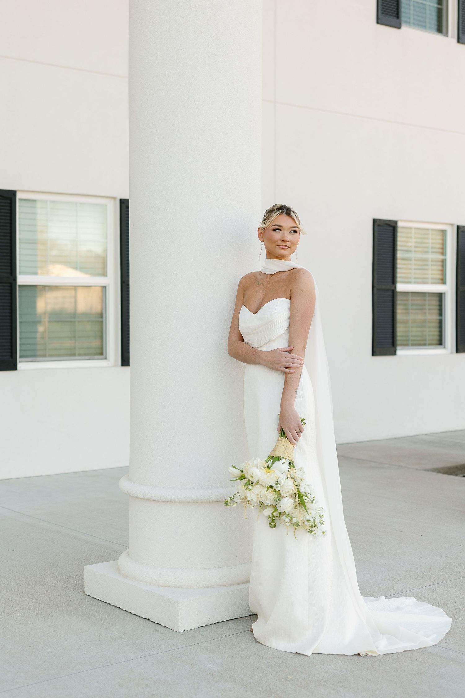 An elegant bride in a white strapless gown with a flowing veil poses beside a white column holding a cream floral bouquet.