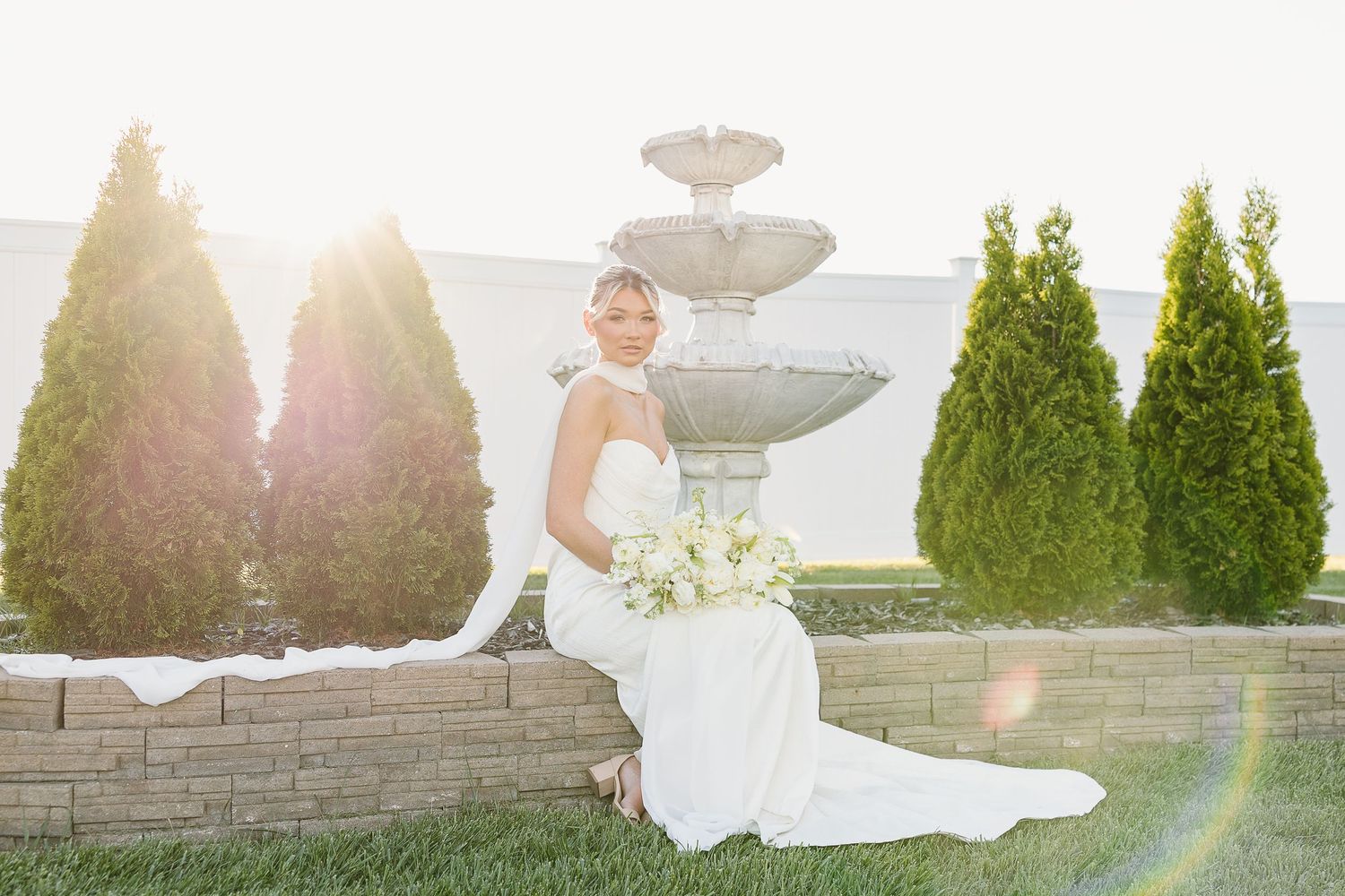 A bride in a white gown sits elegantly by a tiered fountain in a garden setting with evergreen trees during sunset.