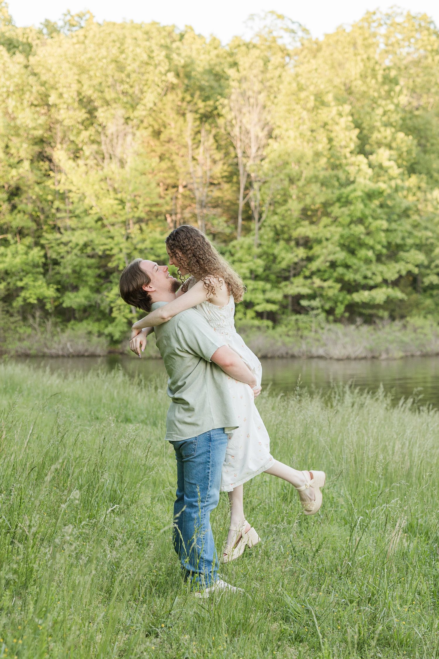 A couple in casual attire embrace in a sunlit meadow by a pond with trees in the background.