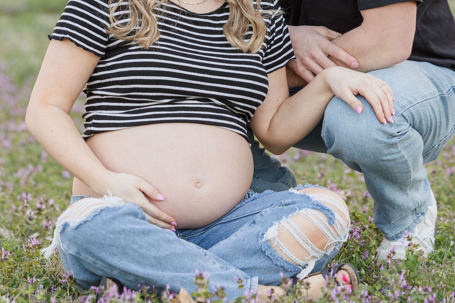 Pregnant woman in striped shirt and ripped jeans sits on grass while someone kneels beside her.