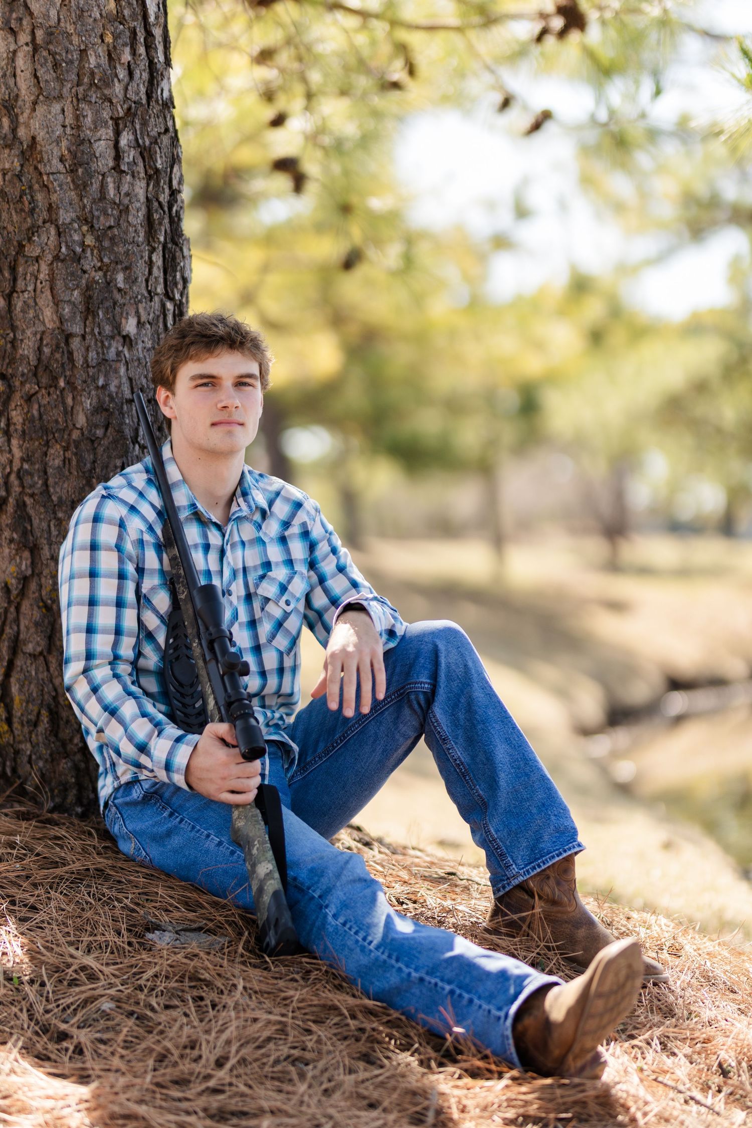 Person in blue plaid shirt sitting against tree trunk holding camera outdoors.